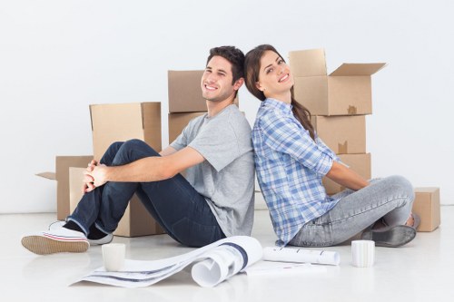 A young man and woman sit back-to-back on a clean, white floor in a bright room, surrounded by various cardboard boxes, some open and others closed, indicating packing for a house move in Maida Vale, London. The man is dressed in a casual grey t-shirt and dark jeans, with short dark hair, smiling as he looks slightly to the side. The woman wears a blue checked shirt and light grey jeans, with long dark hair, gazing upwards with a relaxed expression. In front of them are packing materials, including a rolled-up sheet of paper, a few white mugs, and some additional packing papers, suggesting an organized household packing process. The background is plain with no furniture or décor visible, emphasizing the activity of loading or preparing belongings for a move. This scene illustrates key aspects of domestic removals services offered by Maida Vale Man with Van, highlighting packing and logistical preparation within a typical residential environment.