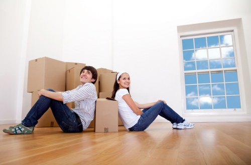 A cheerful young man and woman sit on a polished wooden floor inside a bright, empty room with white walls, surrounded by a variety of cardboard boxes of different sizes, some stacked behind them and others arranged around their legs. The man, wearing a light blue and white striped shirt, jeans, and grey socks with patterns, leans back against the boxes with a relaxed expression. The woman, dressed in a white top, jeans, and white sneakers, sits with her legs extended and crossed at the ankles, smiling at the camera. Large window with multiple panes is visible on the right side, revealing a clear blue sky with a few clouds outside, suggesting daytime. The scene appears to be part of a house move or packing process in an area of Maida Vale, London, consistent with professional removals or house relocation services known in the area, as indicated by the simple, uncluttered interior and natural lighting. The environment is tidy, emphasizing readiness for moving activities handled by a local removal company such as Maida Vale Man with Van.
