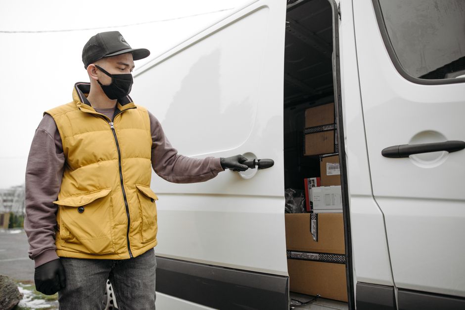 A man with dark hair and medium skin tone, wearing a burgundy polo shirt and a grey cap, is standing beside a white delivery van with the words 'DELIVERY ALWAYS ON TIME' written on the side. He is smi