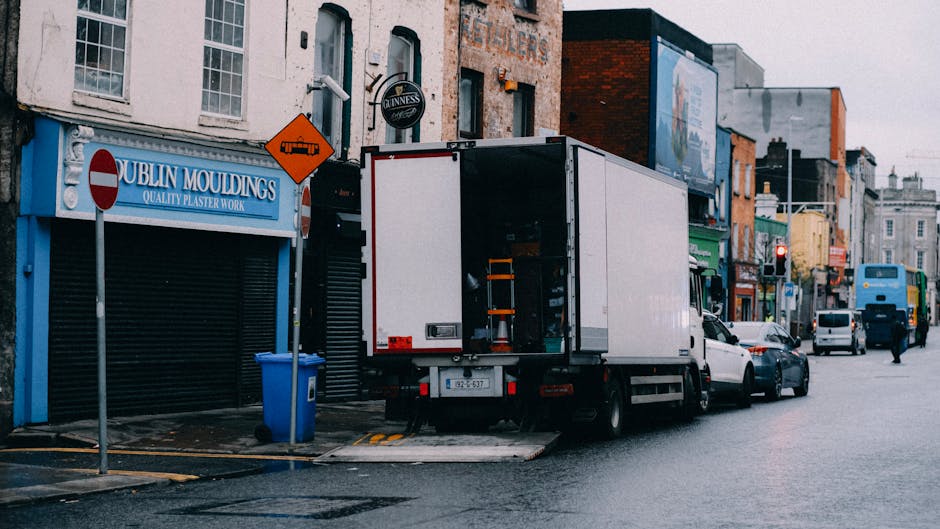 A large white residential moving vehicle parked on a street with its side door closed. In front of the vehicle, a blue recycling bin is positioned on the pavement, with some dirt visible on its surfac
