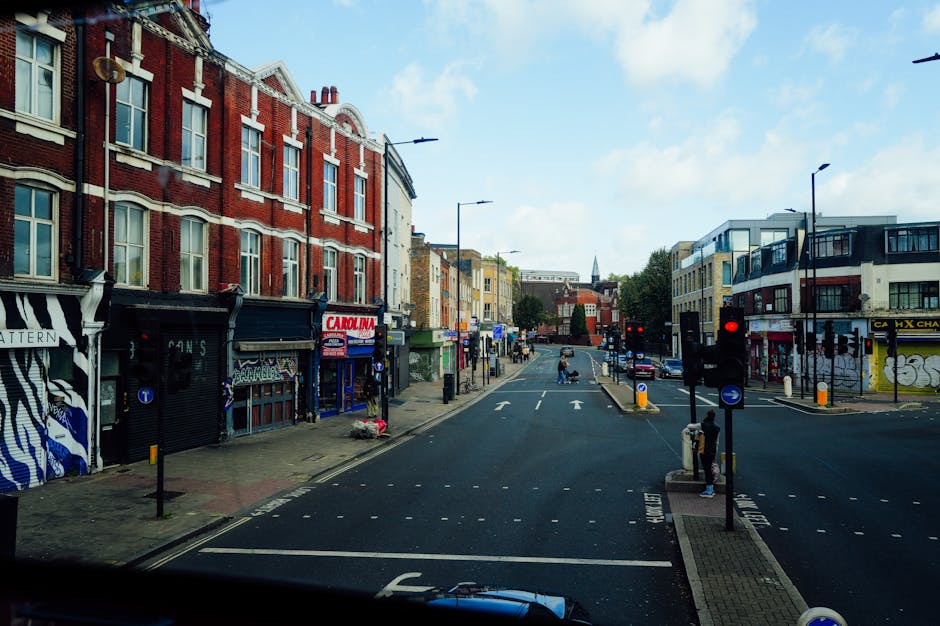 A daytime street scene showing a busy urban area with a variety of commercial and residential buildings lining both sides of the road, which is part of a household removal process. To the left, there 