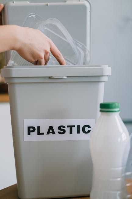 A person disposing of plastic waste into a grey labeled recycling bin with a white label reading 'PLASTIC'. The bin is situated on a wooden surface, with a partially visible plastic bottle placed near