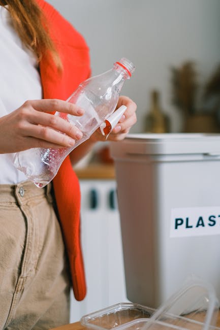 A person disposing of plastic waste into a grey labeled recycling bin with a white label reading 'PLASTIC'. The bin is situated on a wooden surface, with a partially visible plastic bottle placed near